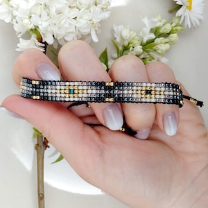 May include: A multi-colored beaded bracelet with black, white, gold, and pink beads. The bracelet is tied with a black cord.