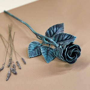 May include: A teal blue metal rose with leaves, lying on a brown surface. A bundle of dried lavender is in the lower left corner of the image.