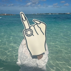 May include: A white foam hand with a black outline and a diamond ring on the index finger. The hand is pointing up and is held in front of a person wearing a white dress. The background is a blue sky and turquoise water.