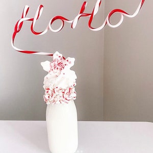 May include: A glass bottle filled with a white milkshake topped with a white whipped cream and crushed candy canes. A red and white striped candy cane straw is bent into the shape of the word "Ho Ho" above the bottle.