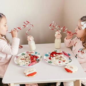 May include: Two young girls wearing pink pajamas sit at a white table with Santa Claus themed plates and cookies. They are drinking milk from glass bottles with red and white striped straws that spell "Holly" and "Jingle".