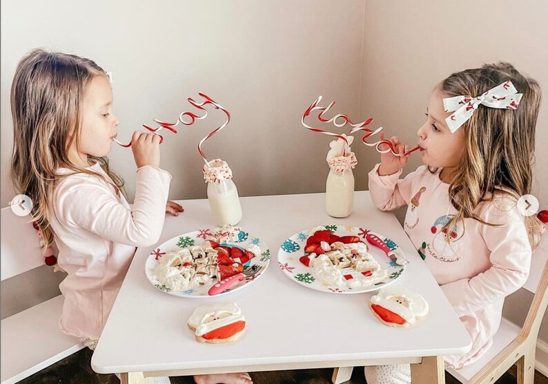 May include: Two young girls wearing pink pajamas sit at a white table with Santa Claus themed plates and cookies. They are drinking milk from glass bottles with red and white striped straws that spell "Holly" and "Jingle".