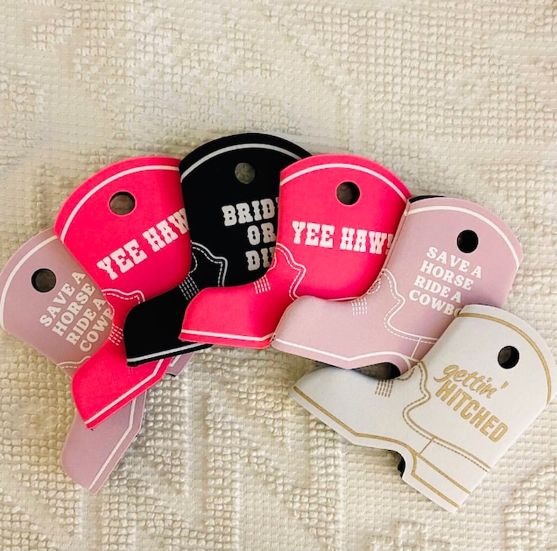 May include: Five pink, black, and white boot-shaped koozies with different sayings. The sayings are "Save a Horse Ride a Cowboy", "Yee Haw", "Bride or Die", "Yee Haw", and "Gettin' Hitched".