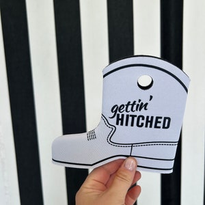 May include: White boot-shaped can cooler with black stitching and the text "gettin' hitched" in black.