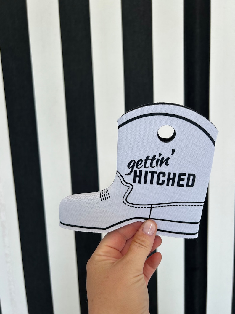 May include: White boot-shaped can cooler with black stitching and the text "gettin' hitched" in black.