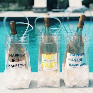May include: Three clear plastic wine totes with handles, each containing a bottle of white wine and ice. The totes have different text printed on them: "Happier in the Hamptons", "Out East", and "Happier in the Hamptons".