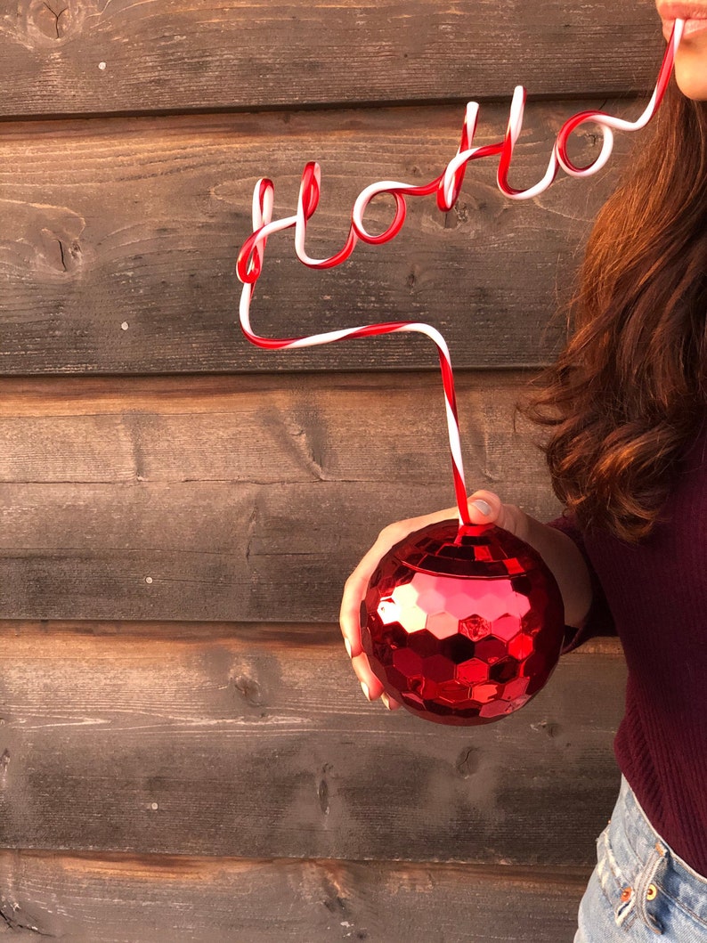 May include: A red and white striped drinking straw bent into the word "Ho Ho" with a red disco ball ornament hanging from the straw.