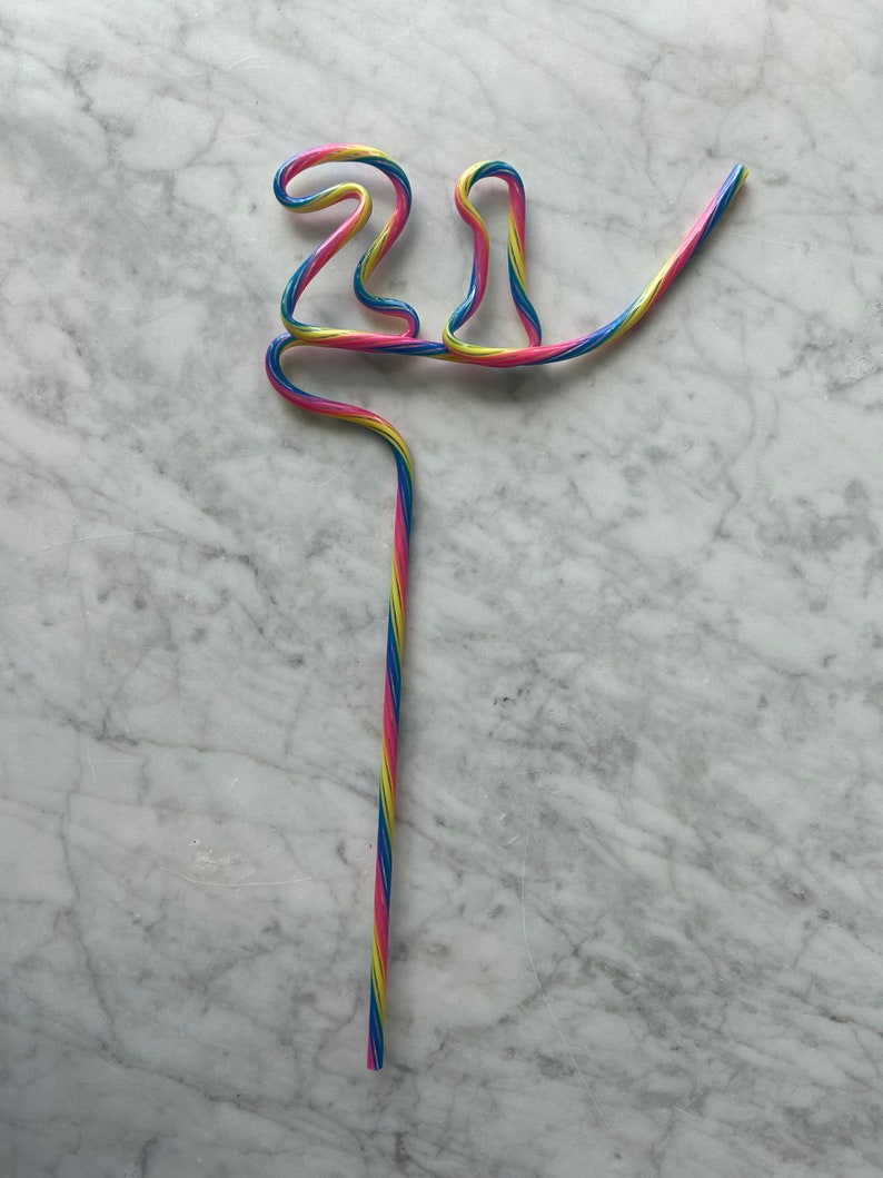 May include: A colorful, striped drinking straw bent into the shape of the number 21. The straw is pink, blue, and yellow.