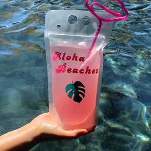 May include: A clear plastic pouch with a pink drink inside. The pouch has a pink straw with a lip-shaped top and the words "Aloha Beaches" printed on the front. A green monstera leaf is also printed on the front of the pouch.