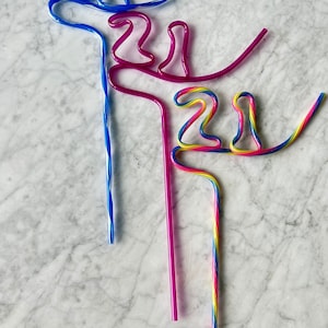 May include: Three colorful bendy straws shaped into the number 21. The straws are blue, pink, and rainbow.