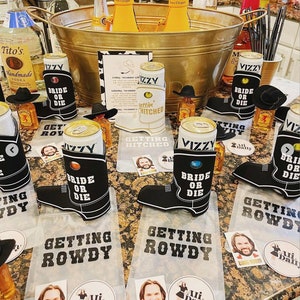 May include: A table with a variety of drinks and snacks for a party. There are several cans of beer with black and white boot-shaped koozies that say "Bride or Die" and "Getting Rowdy". There are also several small bottles of liquor and a large metal tub filled with ice and champagne bottles. The table is decorated with several small signs that say "Getting Hitched" and "Getting Rowdy".