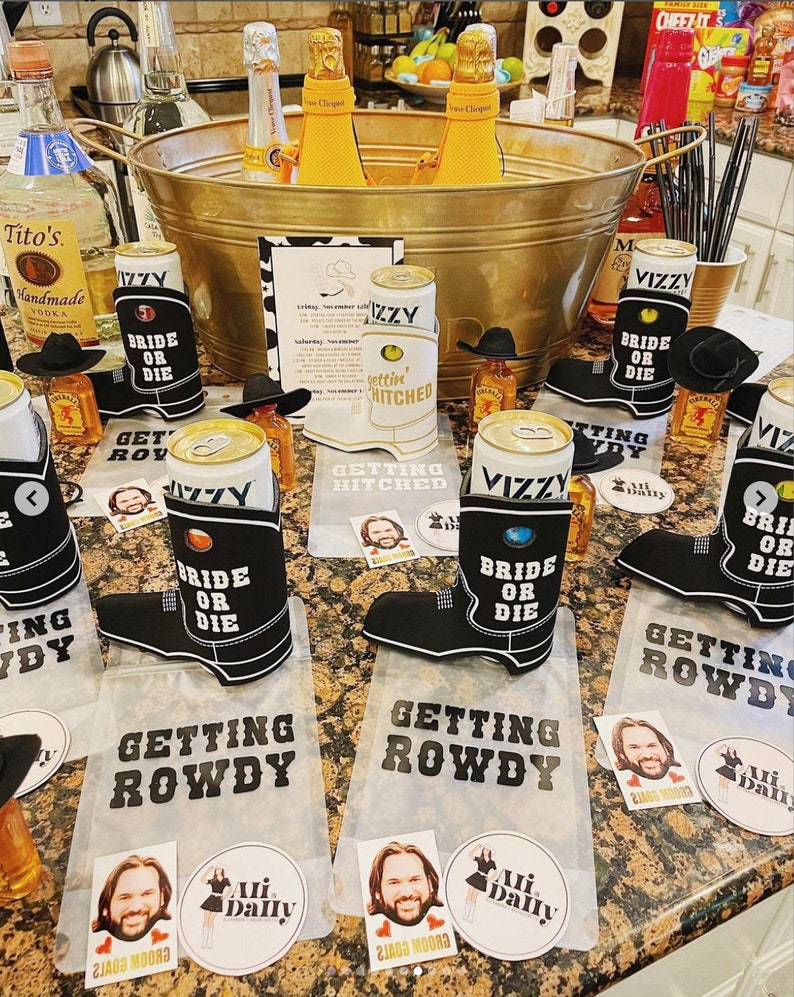 May include: A table with a variety of drinks and snacks for a party. There are several cans of beer with black and white boot-shaped koozies that say "Bride or Die" and "Getting Rowdy". There are also several small bottles of liquor and a large metal tub filled with ice and champagne bottles. The table is decorated with several small signs that say "Getting Hitched" and "Getting Rowdy".