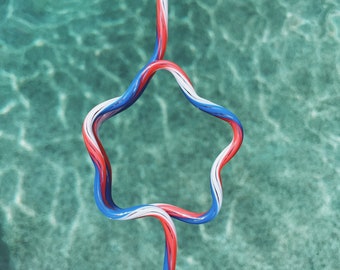 May include: A red, white, and blue striped drinking straw bent into the shape of a star. The straw is floating in a pool of blue water.