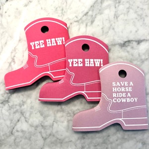 K&ouml;nnte beinhalten: Drei rosa Cowboy-Stiefel aus Schaumstoff als Dekoration. Die beiden oberen haben den Text "Yee Haw!" aufgedruckt. Die untere Dekoration hat den Text "Save a horse, ride a cowboy" aufgedruckt.