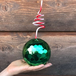 May include: A green, iridescent, round, disco ball-shaped cup with a red and white striped straw. The cup is being held in a hand.