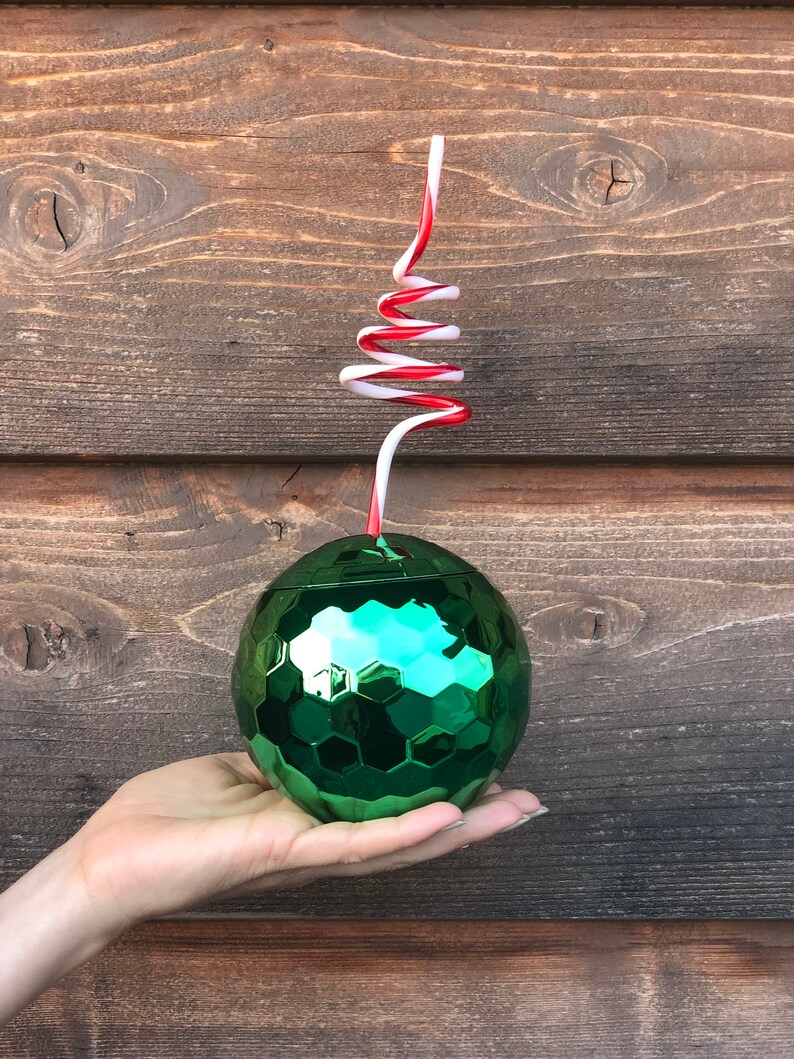 May include: A green, iridescent, round, disco ball-shaped cup with a red and white striped straw. The cup is being held in a hand.
