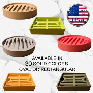 May include: A collection of oval and rectangular soap dishes in various solid colors including tan, lime green, red, orange, and olive. The dishes feature slotted drainage designs. Text reads: MADE IN USA, AVAILABLE IN 30 SOLID COLORS, OVAL OR RECTANGULAR.