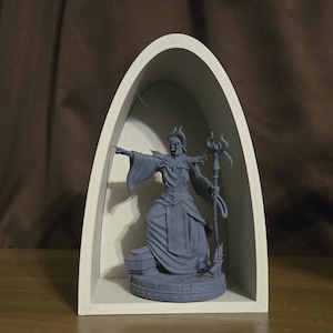 May include: A gray figurine of a robed figure with a staff, inside a light-colored arched display case. The figure is pointing with one hand. The figurine is on a round base. The display case is set against a brown background.