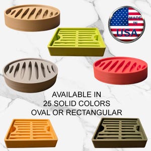 May include: An assortment of colorful, slotted soap dishes in oval and rectangular shapes. The image shows dishes in shades of tan, green, beige, red, orange, and olive green. Text on the image reads "AVAILABLE IN 25 SOLID COLORS OVAL OR RECTANGULAR" and a "MADE IN USA" graphic.