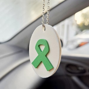 May include: A white oval-shaped car charm with a green ribbon design. The charm is suspended from a silver chain. The ribbon is a symbol of awareness and support.