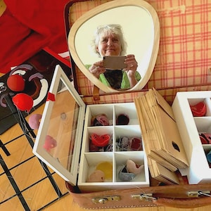 May include: A vintage brown suitcase containing two white wooden organizers with multiple compartments filled with small, colorful, rounded objects. A teardrop-shaped mirror is attached to the interior lid, and a stack of brown paper envelopes sits between the organizers.