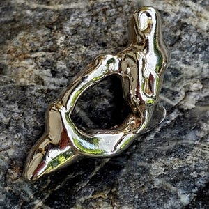 May include: A polished silver pendant with an abstract, organic design. The pendant has a flowing, curved shape with a large central opening. The reflective surface catches light, highlighting the form's contours. The pendant is displayed on a textured gray stone surface.