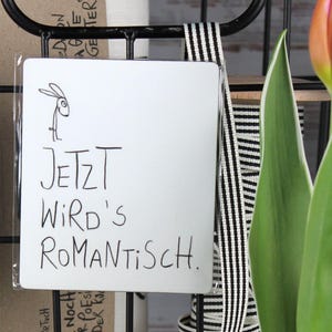 Könnte beinhalten: Ein schwarzer Drahtkorb enthält ein weißes Schild mit dem deutschen Text "JETZT WIRD'S ROMANTISCH" und einer kleinen Hasenzeichnung. Orange und rosa Tulpenknospen mit grünen Blättern sind im Vordergrund, was auf ein Frühlingsmotiv hindeutet.