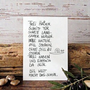 Könnte beinhalten: Eine handgeschriebene Notiz mit deutschem Text und einer Schneckenzeichnung auf weißem Papier. Vor einem rustikalen Holzuntergrund, mit Oliven und Steinen, die eine natürliche Ästhetik erzeugen.