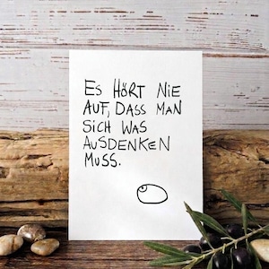 Op de afbeelding: Een witte kaart met zwarte handgeschreven tekst in het Duits: "Es hört nie auf, dass man sich was ausdenken muss." Een eenvoudige oogtekening staat eronder. De kaart ligt op een houten oppervlak met olijven en bladeren.