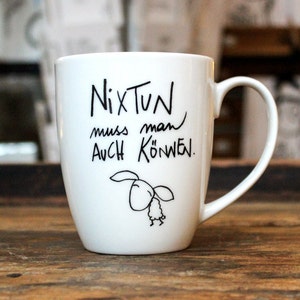 Könnte beinhalten: Weiße Keramiktasse mit einer schwarzen handschriftlichen Botschaft, die "Nix tun muss man auch können." lautet. Die Tasse hat eine schwarz-weiße Illustration eines Schafes mit einem Blatt im Maul.