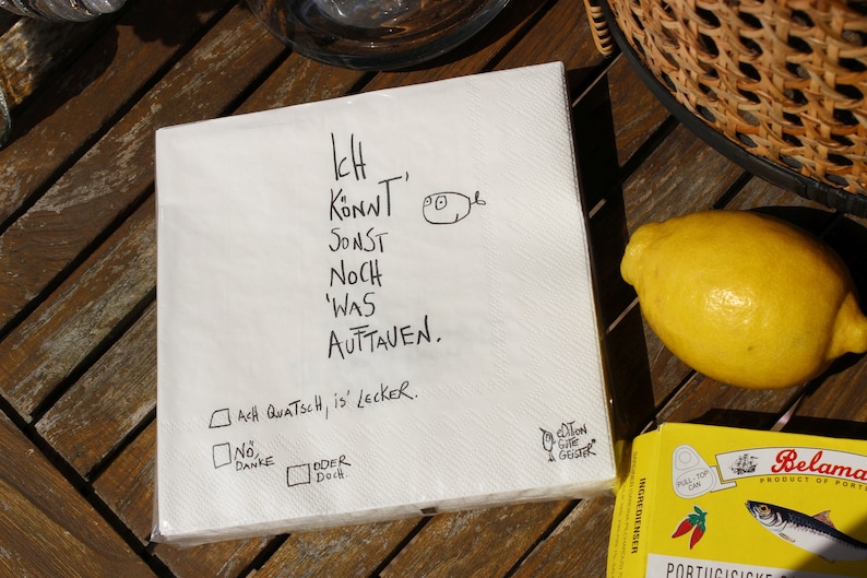 May include: A white paper napkin with black handwritten text that says "Ich Konnt Sonst Noch Was Auttaven. Ach Quatsch, is lecker. No Danke Oder Doch." There are two checkboxes, one with a checkmark and one without. The napkin is on a wooden table with a lemon and a can of sardines in the background.