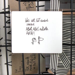 Könnte beinhalten: Ein schwarzer Drahtkorb mit einem weißen Schild mit der Aufschrift "Was wir gut können: streiten. Aber alles andere auch!" in schwarzer Handschrift. Das Schild hat zwei kleine Cartoon-Zeichnungen. Schwarz-weiß gestreiftes Band hängt vom Korb.