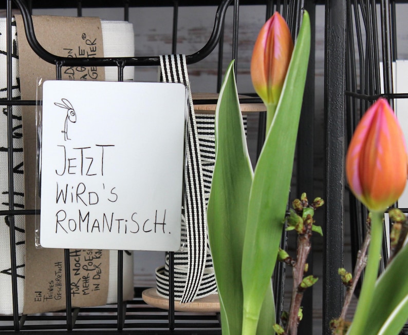 Könnte beinhalten: Ein schwarzer Drahtkorb enthält ein weißes Schild mit dem deutschen Text "JETZT WIRD'S ROMANTISCH" und einer kleinen Hasenzeichnung. Orange und rosa Tulpenknospen mit grünen Blättern sind im Vordergrund, was auf ein Frühlingsmotiv hindeutet.