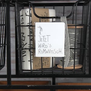 Puede incluir: Una cesta de alambre negra llena de materiales de manualidades. Contiene rollos de papel, una bobina de cinta a rayas blancas y negras y un letrero blanco que dice "JETZT WIRD'S ROMANTISCH."