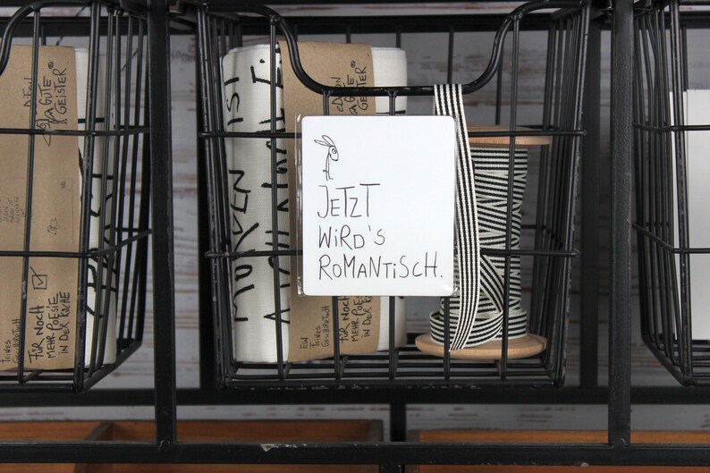 Puede incluir: Una cesta de alambre negra llena de materiales de manualidades. Contiene rollos de papel, una bobina de cinta a rayas blancas y negras y un letrero blanco que dice "JETZT WIRD'S ROMANTISCH."