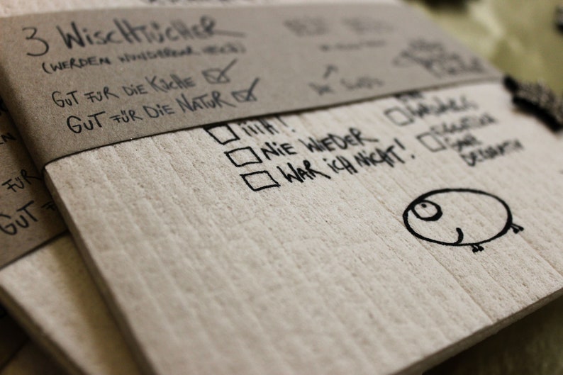 May include: A close-up of a brown paper package with the text "3 WiscHTUCHER (WERDEN WHitech GUT FUR DIE KICHE✓ GUT F&Uuml;R DIE NATURAㄧˋˋˋ" written on it. The package is folded over and has a white check mark next to the text "GUT FUR DIE KICHE". The package is also labeled with the text "NIE WIEDER" and "WAR ICH NICHT!" in black marker.