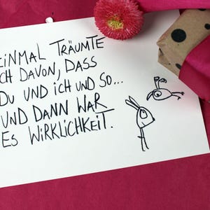 May include: A white card with handwritten German text and simple illustrations of a rabbit and bird. A wrapped gift with a polka dot pattern and a pink ribbon sits next to the card on a pink surface.
