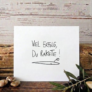 May include: A white card with the handwritten text "Viel Erfolg, Du Rakete!" and a small rocket illustration. The card rests on a wooden surface, with olives and olive branches in the foreground.