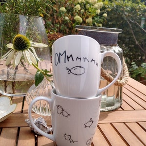 Peut inclure: Deux tasses en céramique blanche empilées l'une sur l'autre sur une table en bois. La tasse du haut porte le mot "OMMMMM" écrit en noir. La tasse du bas a un dessin au marqueur noir d'un oiseau, d'un chat et d'un mouton.