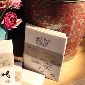 May include: Three white dishcloths with a brown label that reads "3 Wischt&uuml;cher (werden wieder weich)  Got war die Natur...  Die sind's...  Harthackig  Eigentlich  ganz  dekorativ" and a black and white logo that reads "edition gute geister". The label also has a check box with the text "Nie wieder war ich nicht!" and another check box with the text "Stor auf die Kiche". The dishcloths are stacked on top of each other and have a white background with black text that reads "Jeder Fleck erz&auml;hlt eine Geschichte..."