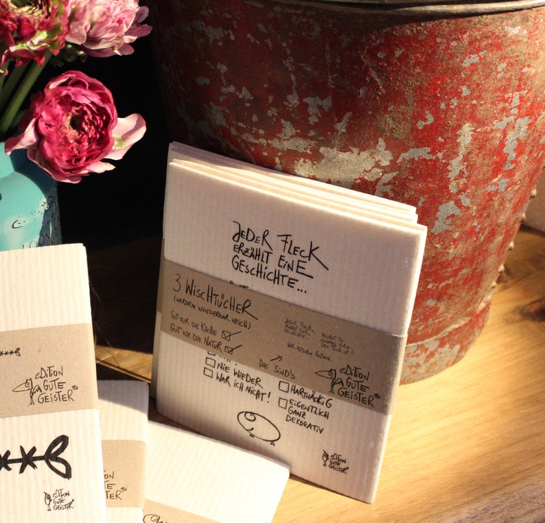 May include: Three white dishcloths with a brown label that reads "3 Wischt&uuml;cher (werden wieder weich)  Got war die Natur...  Die sind's...  Harthackig  Eigentlich  ganz  dekorativ" and a black and white logo that reads "edition gute geister". The label also has a check box with the text "Nie wieder war ich nicht!" and another check box with the text "Stor auf die Kiche". The dishcloths are stacked on top of each other and have a white background with black text that reads "Jeder Fleck erz&auml;hlt eine Geschichte..."