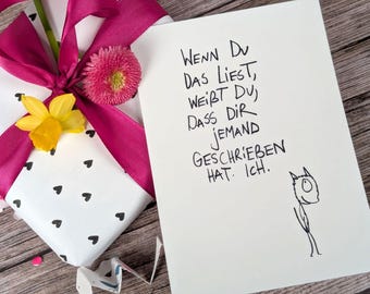 May include: A wrapped gift with a white background and black heart pattern, adorned with a magenta ribbon and a yellow daffodil. A card with handwritten German text and a simple drawing is placed next to the gift.