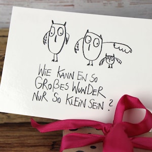 Puede incluir: Una tarjeta blanca con una ilustración dibujada a mano de tres búhos. La tarjeta tiene el texto alemán "Wie kann ein so großes Wunder nur so klein sein?" Una cinta rosa está atada alrededor de un regalo.