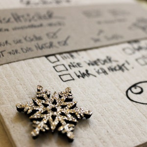 May include: A white card with handwritten text in black ink. The text reads "3 Wisemen", "Got for the kids", "Gut for die Natur", "Nie Wetek", "War ich nicht!" and "Jedes Jahr". A silver snowflake ornament is on the card.