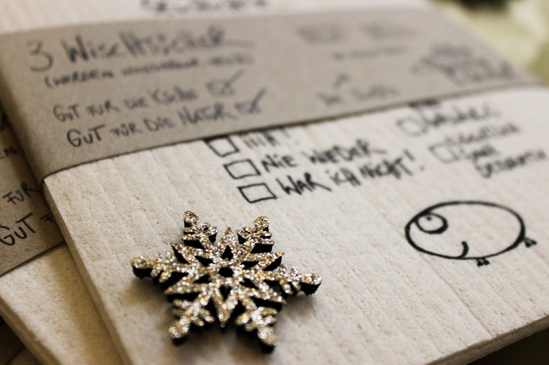May include: A white card with handwritten text in black ink. The text reads "3 Wisemen", "Got for the kids", "Gut for die Natur", "Nie Wetek", "War ich nicht!" and "Jedes Jahr". A silver snowflake ornament is on the card.