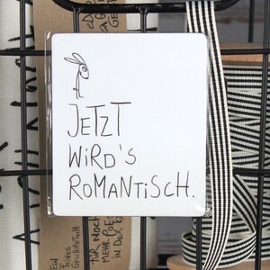 Puede incluir: Un letrero rectangular blanco con el texto alem&aacute;n "JETZT WIRD'S ROMANTISCH" y una ilustraci&oacute;n de un conejo. El letrero est&aacute; en una cesta de alambre negra con otros art&iacute;culos.