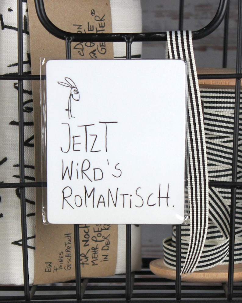 Puede incluir: Un letrero rectangular blanco con el texto alem&aacute;n "JETZT WIRD'S ROMANTISCH" y una ilustraci&oacute;n de un conejo. El letrero est&aacute; en una cesta de alambre negra con otros art&iacute;culos.