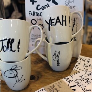 Könnte beinhalten: Weiße Keramikbecher mit schwarzem handgeschriebenem Text und skurrilen Zeichnungen. Auf einem Becher steht "YEAH!" und auf einem anderen "Toll!". Einige Becher zeigen eine Kaninchen-Illustration. Die Becher sind gestapelt.