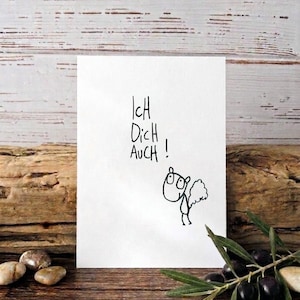 Könnte beinhalten: Eine weiße Karte mit dem handgeschriebenen deutschen Text "Ich Dich Auch!" und einer kleinen Cartoon-Zeichnung. Die Karte ist vor einem rustikalen Holzgrund, mit Oliven und Steinen.