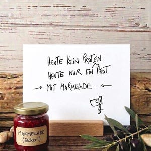 Könnte beinhalten: Ein Glas rote Marmelade mit goldenem Deckel und einem Etikett mit der Aufschrift "Marmelade (lecker!)". Eine handschriftliche Notiz lautet "HEUTE KEIN PROTEIN. HEUTE NUR EIN PROT MIT MARMELADE." Eine Zeichnung einer Person befindet sich auf der Notiz. Oliven und ein Holzständer sind ebenfalls vorhanden.
